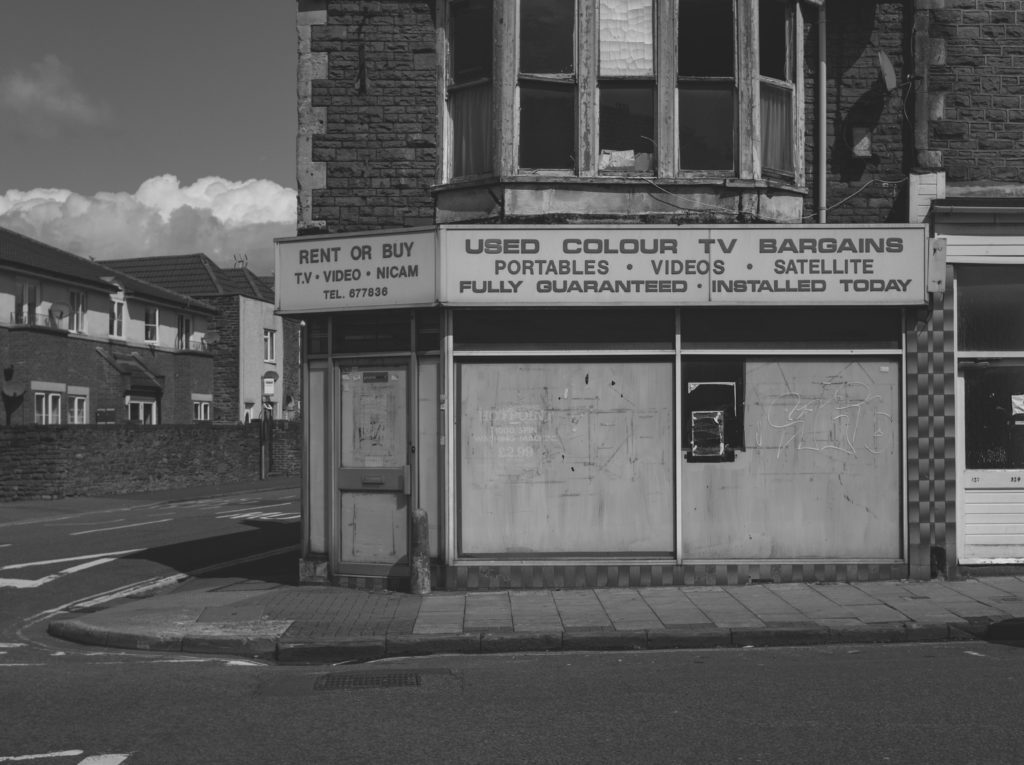 Black and white photo of a closed-down corner shop advertising used TVs.