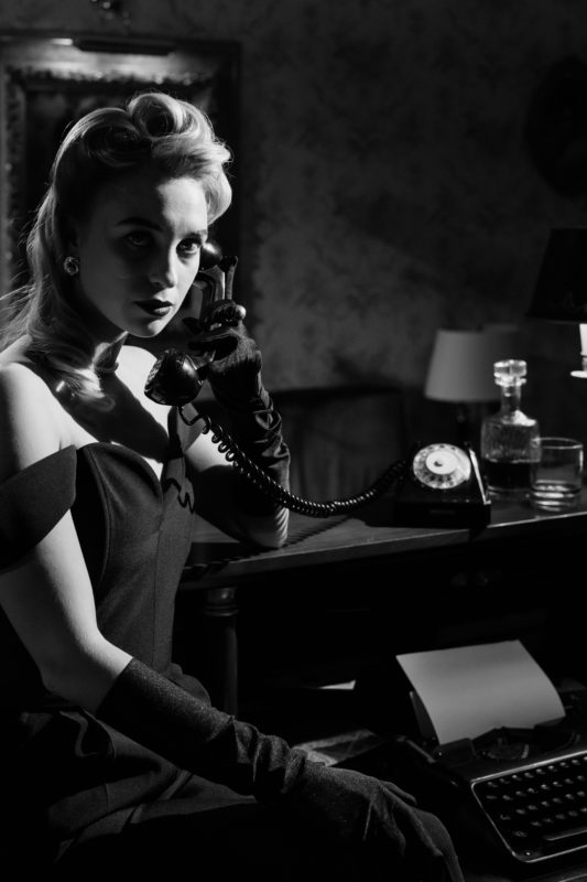 A high-contrast monochrome photograph of a woman in early twentieth century formalwear answering a phone.