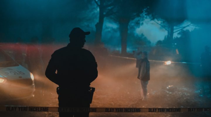 Several people standing in the woods at night behind crime scene tape, a cool blue backlight lancing through mist while the foreground is lit by warmer car headlights.